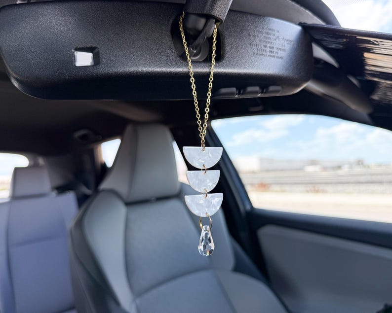 May include: A hanging car ornament featuring a gold-toned chain connected to three white, semi-circular acrylic pieces with a marbled texture, finished with a clear, faceted teardrop-shaped crystal pendant suspended at the bottom of the chain.