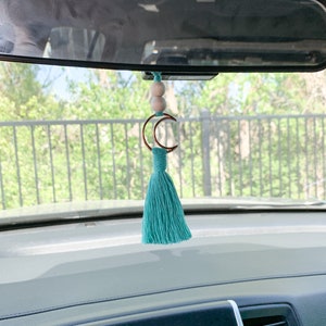 May include: A car rearview mirror hanging ornament featuring two light-colored wooden beads, a copper-toned crescent moon charm, and a teal-colored fiber tassel suspended from a matching teal cord.