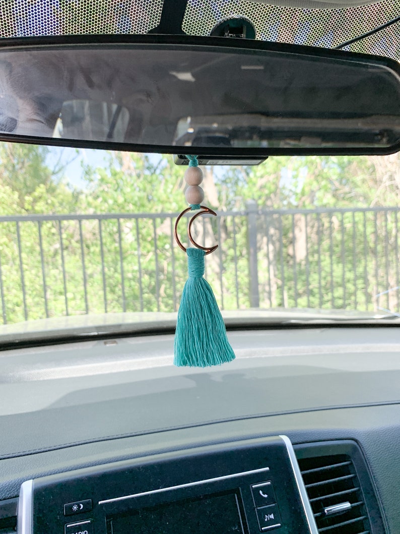 May include: A car rearview mirror hanging ornament featuring two light-colored wooden beads, a copper-toned crescent moon charm, and a teal-colored fiber tassel suspended from a matching teal cord.