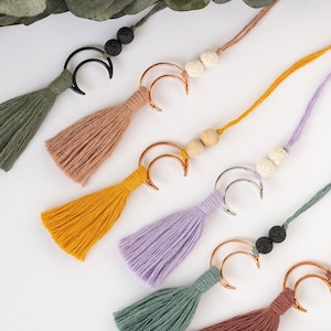 May include: A collection of six decorative tassels, each featuring a crescent moon charm in black, gold, or silver metal. The tassels are made of colored thread in shades of olive, tan, mustard, lavender, and rust, accented with two round beads.
