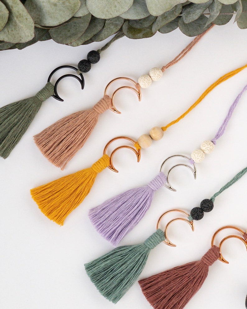 May include: A collection of six decorative tassels, each featuring a crescent moon charm in black, gold, or silver metal. The tassels are made of colored thread in shades of olive, tan, mustard, lavender, and rust, accented with two round beads.
