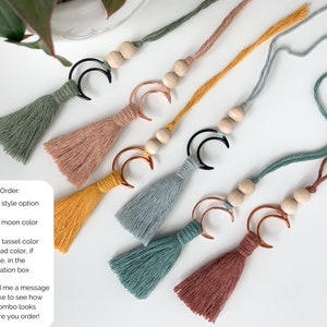 May include: Five hanging ornaments featuring cotton tassels in sage green, dusty rose, mustard yellow, slate blue, and terracotta. Each ornament includes two natural wooden beads and a crescent moon charm in either black or copper metal finishes.