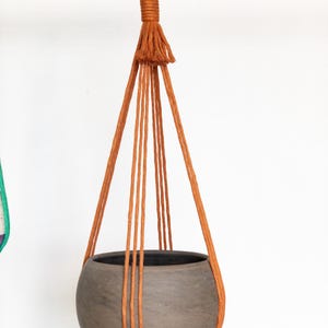 May include: A hanging planter with a round, gray ceramic pot suspended by rust-colored macrame cords. The cords are attached to a metal ring at the top, creating a stylish and earthy aesthetic. The planter is designed for indoor or outdoor use.