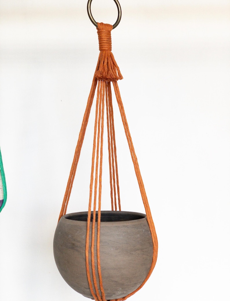 May include: A hanging planter with a round, gray ceramic pot suspended by rust-colored macrame cords. The cords are attached to a metal ring at the top, creating a stylish and earthy aesthetic. The planter is designed for indoor or outdoor use.