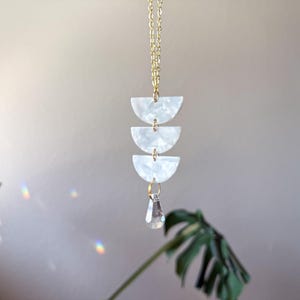 May include: A gold-toned chain necklace featuring three stacked white semi-circular pieces with a pearlescent finish, connected by small gold rings, and finished with a single clear, faceted teardrop-shaped crystal pendant hanging at the bottom.