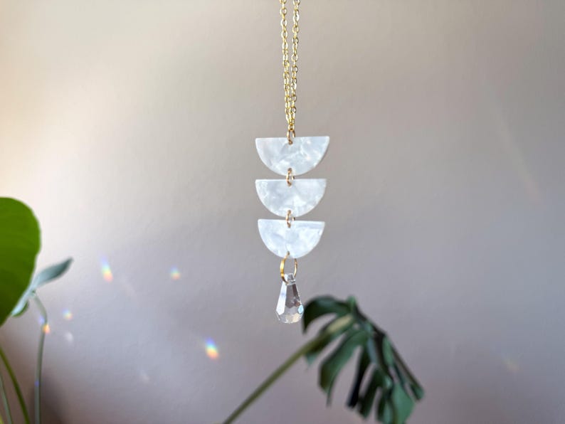 May include: A gold-toned chain necklace featuring three stacked white semi-circular pieces with a pearlescent finish, connected by small gold rings, and finished with a single clear, faceted teardrop-shaped crystal pendant hanging at the bottom.