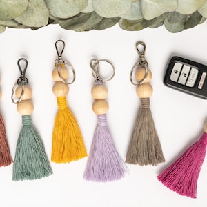 May include: Six keychains featuring two natural wooden beads and a colorful cotton tassel in shades of rust, sage green, mustard yellow, lavender, taupe, and magenta, each attached to a metal lobster clasp and a split ring, with one car key fob.