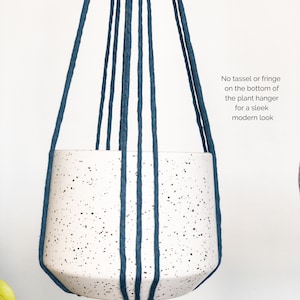 May include: A white speckled ceramic plant pot suspended by dark blue rope. The text on the image reads: "No tassel or fringe on the bottom of the plant hanger for a sleek modern look."