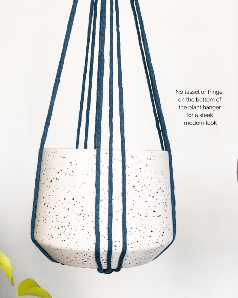 May include: A white speckled ceramic plant pot suspended by dark blue rope. The text on the image reads: "No tassel or fringe on the bottom of the plant hanger for a sleek modern look."