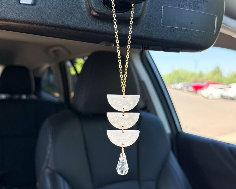 May include: A car rearview mirror hanging ornament featuring a gold-toned chain connected to three stacked white marbled acrylic semi-circle shapes, finished with a clear, faceted, teardrop-shaped crystal pendant dangling at the bottom.