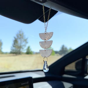 May include: A car rearview mirror hanging ornament featuring three stacked white semi-circular pieces with a pearlescent finish, suspended by a gold-toned metal chain and finished with a clear, faceted, teardrop-shaped crystal pendant at the bottom.