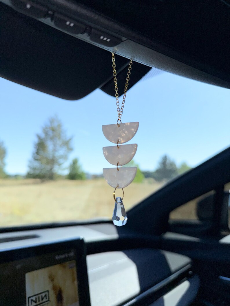 May include: A car rearview mirror hanging ornament featuring three stacked white semi-circular pieces with a pearlescent finish, suspended by a gold-toned metal chain and finished with a clear, faceted, teardrop-shaped crystal pendant at the bottom.