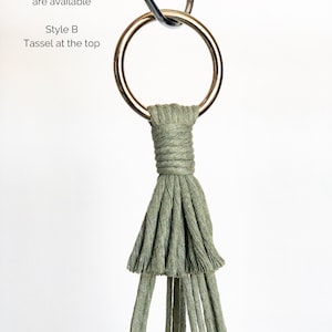 May include: A macrame plant hanger in a sage green color, featuring a metal ring for hanging. The design includes a wrapped section and a tassel at the bottom. Text in the image reads: "2 Style Options are available" and "Style B Tassel at the top."