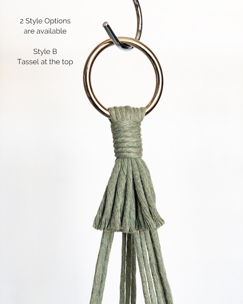 May include: A macrame plant hanger in a sage green color, featuring a metal ring for hanging. The design includes a wrapped section and a tassel at the bottom. Text in the image reads: "2 Style Options are available" and "Style B Tassel at the top."