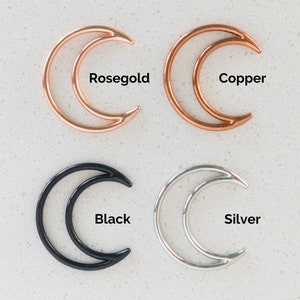 May include: Four crescent moon-shaped metal charms arranged in a grid. The top left is rosegold, the top right is copper, the bottom left is black, and the bottom right is silver. Each charm is labeled with its respective color name in black text.