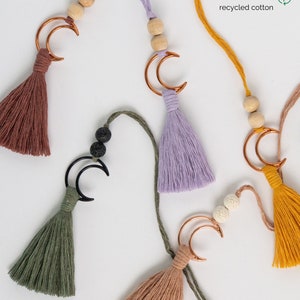 May include: Five hanging ornaments featuring cotton tassels in rust, lavender, olive green, and mustard yellow. Each includes a crescent moon charm, wooden or porous beads, and a cord. Text reads: Sustainably made with soft 100% recycled cotton.