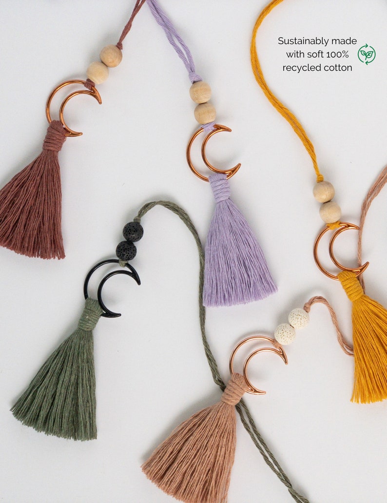 May include: Five hanging ornaments featuring cotton tassels in rust, lavender, olive green, and mustard yellow. Each includes a crescent moon charm, wooden or porous beads, and a cord. Text reads: Sustainably made with soft 100% recycled cotton.
