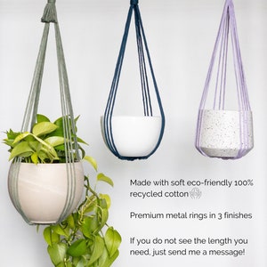 May include: Three hanging planters with metal rings, each suspended by woven cords. One holds a plant with green leaves, another a white pot, and the third a speckled pot. The planters are in shades of green, blue, and purple.