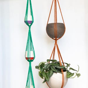 May include: Macrame plant hangers in green and brown, suspended from black metal rings. The green hanger holds three small pots with red and white speckled designs. The brown hanger supports a gray pot and a white pot with trailing green plants.