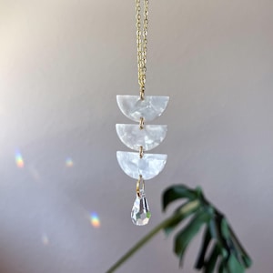May include: A gold chain necklace featuring three stacked white semi-circular acrylic pieces with a pearlescent finish. The pendant is completed by a single clear, faceted teardrop-shaped crystal charm hanging from the bottom of the stack.