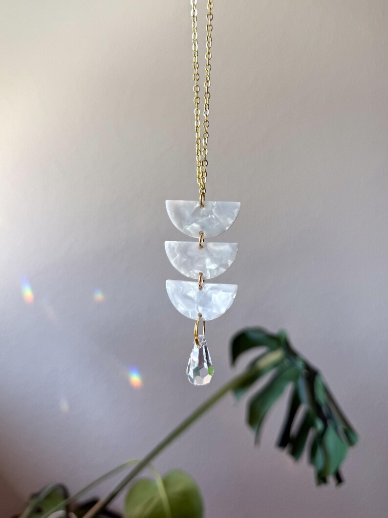 May include: A gold chain necklace featuring three stacked white semi-circular acrylic pieces with a pearlescent finish. The pendant is completed by a single clear, faceted teardrop-shaped crystal charm hanging from the bottom of the stack.