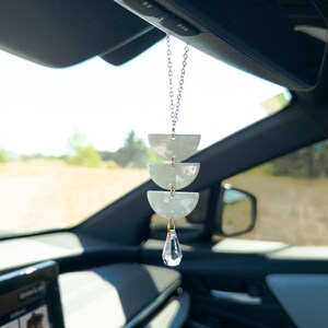 May include: A car hanging ornament featuring three stacked white semi-circle pieces suspended by a silver-toned chain, finished with a clear, faceted crystal drop at the bottom. The ornament hangs from a vehicle rearview mirror.