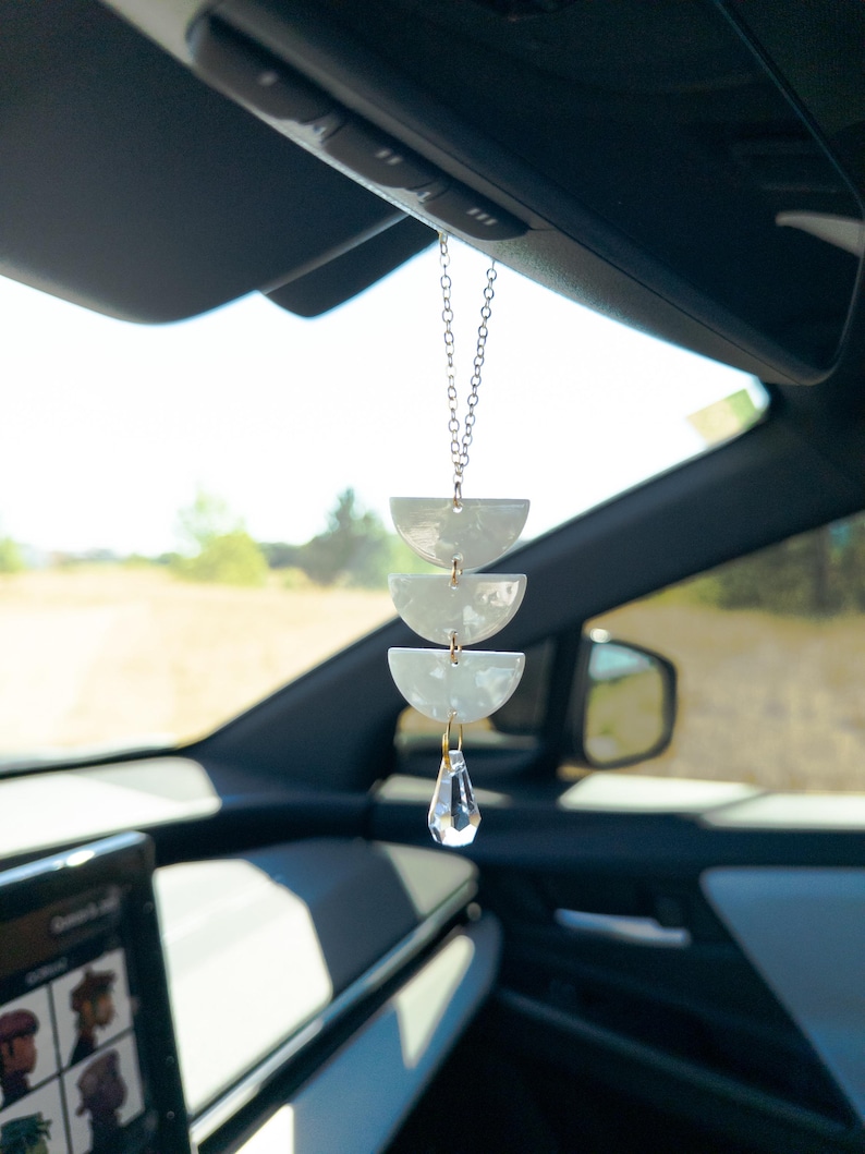 May include: A car hanging ornament featuring three stacked white semi-circle pieces suspended by a silver-toned chain, finished with a clear, faceted crystal drop at the bottom. The ornament hangs from a vehicle rearview mirror.