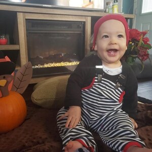 May include: A baby wearing a red and white striped overall with a red hat is sitting on a brown blanket. The baby is smiling and looking at the camera. There is a wooden turkey figurine in the background.
