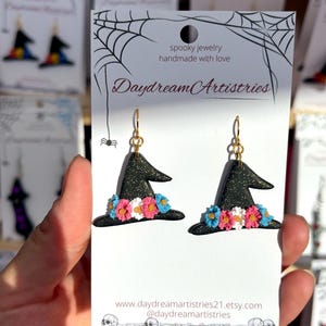 May include: A pair of black witch hat earrings with gold glitter accents and a band of small pink, white, and blue flowers. The earrings are displayed on a white card featuring the text Daydream Artistries, spooky jewelry handmade with love.