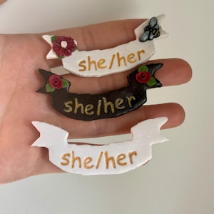 Op de afbeelding: Drie handgemaakte speldjes in de vorm van een banner met de tekst "she/her". Eén is wit met een rode bloem en een bij, één is zwart met een rode roos en de andere is wit. De tekst is in goud.