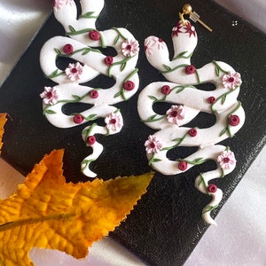 May include: A pair of white snake-shaped earrings adorned with pink flowers and red rose accents. The earrings have gold-colored posts and are decorated with green vines. The earrings are on a black surface.