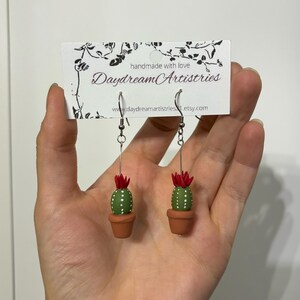 May include: A pair of dangle earrings featuring a small potted cactus with red flowers. The earrings are handmade with love by Daydream Artistries.