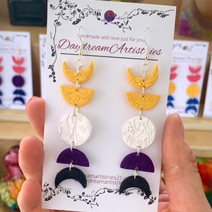 Puede incluir: Pendientes hechos a mano con temática celestial. Cada pendiente presenta una serie de formas coloridas y texturizadas: medias lunas amarillas, semicírculos amarillos, círculos florales blancos, semicírculos morados y medias lunas negras. Los pendientes están suspendidos de ganchos plateados. El texto "Daydream Artistries" es visible.