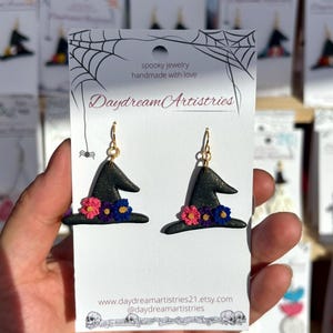 May include: A pair of black witch hat earrings with gold-colored hooks. The hats are decorated with pink, blue, and purple flowers. The earrings are displayed on a white card with the text "spooky jewelry handmade with love" and the shop name "Daydream Artistries".