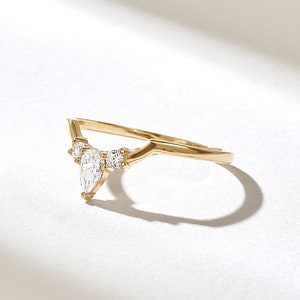 May include: A gold-colored ring with a V-shaped design. The ring features a pear-shaped diamond in the center, flanked by smaller round diamonds. The ring is set against a white background, with a shadow.