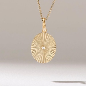 Op de afbeelding: Een goudkleurige hangerketting. De ovale hanger heeft een sunburst-ontwerp met een kleine, ronde, heldere edelsteen in het midden. De ketting is delicaat en ook goudkleurig. De hanger hangt aan een kleine lus.