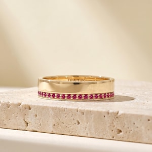 May include: A gold band ring featuring a single row of small, round, vibrant pink stones set along the lower edge. The interior of the band is engraved with the text DIA 14K, indicating the metal composition. The ring has a polished, smooth finish.