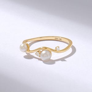 Op de afbeelding: Een 14K gouden ring met een golvend ontwerp, twee witte parels van verschillende groottes en twee kleine, heldere, rondgeslepen diamanten. De binnenkant van de ring is gegraveerd met de tekst DIA 14K, wat de materiaalsamenstelling aangeeft.
