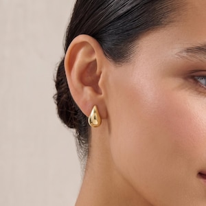 May include: A polished gold earring in the shape of a smooth, rounded teardrop. The earring features a reflective, metallic surface with a curved, bulbous form that tapers to a point at the top, securely fastened to the earlobe with a post.
