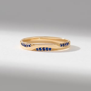 May include: A gold band ring featuring a wave-like design with small, round, deep blue sapphires set along the curved sections. The polished gold metal creates a smooth, continuous surface that contrasts with the textured, stone-set segments.