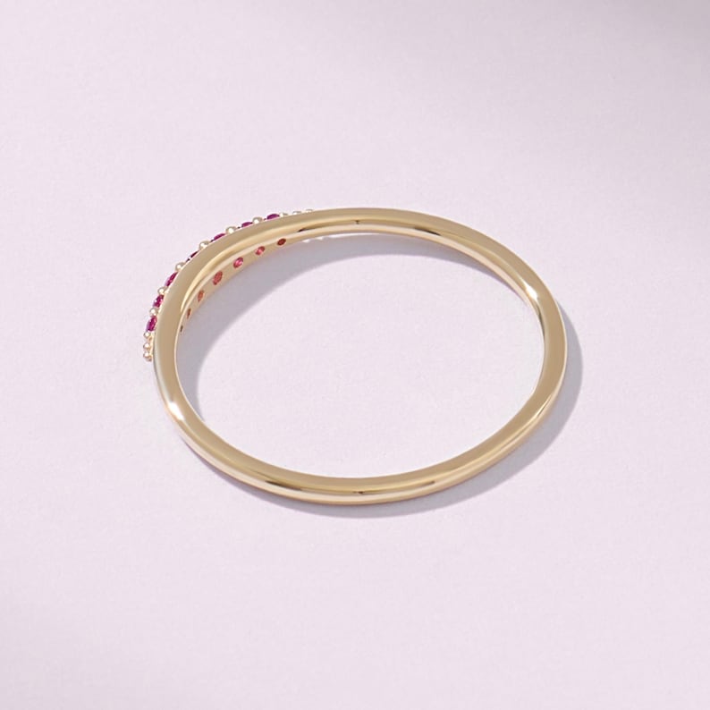 Puede incluir: Un anillo de oro con un dise&ntilde;o curvo. El anillo presenta una fila de peque&ntilde;as piedras preciosas rojas redondas engastadas a lo largo de la curva. El anillo est&aacute; sobre un fondo blanco, resaltando los colores oro y rojo.