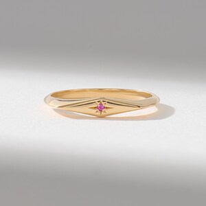 May include: A gold band ring featuring a central marquise-shaped setting with a starburst engraving. A small, round, vibrant pink gemstone is set securely in the center of the starburst, creating a focal point on the polished gold surface.