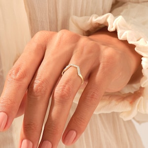 Könnte beinhalten: Ein goldfarbener Ring in V-Form, besetzt mit kleinen, klaren Edelsteinen. Der Ring befindet sich an einem Finger, mit einem weichen, neutralen Hintergrund. Das Design des Rings ist zart und elegant.