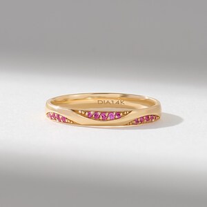 May include: A 14k gold band ring featuring a wave-like design set with small, round, vibrant pink gemstones. The interior of the band is clearly engraved with the text DIA14K, indicating the metal purity and material composition.