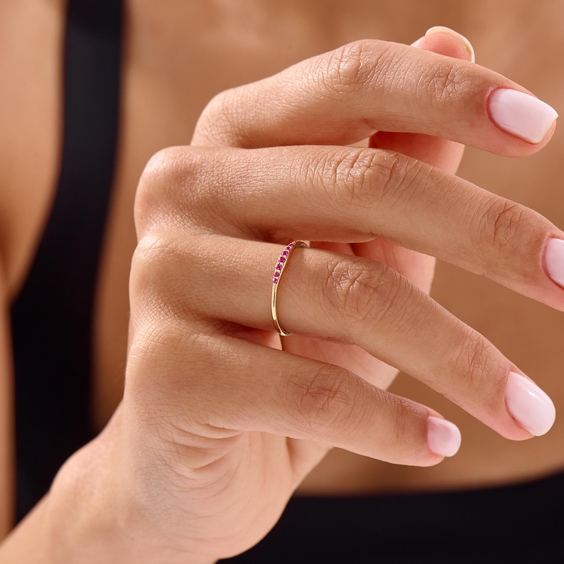 Puede incluir: Un anillo de oro con una fila de peque&ntilde;as piedras preciosas de color rosa brillante. El anillo es delgado y delicado, y las piedras preciosas est&aacute;n espaciadas uniformemente. El anillo se muestra en un dedo.