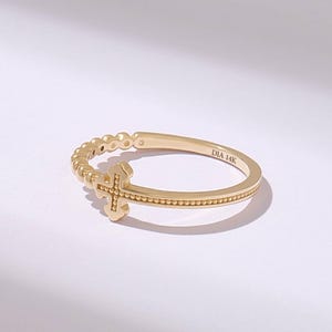 May include: A gold ring featuring a cross design with a beaded texture along the band and the cross itself. The interior of the band is clearly engraved with the text DIA 14K, indicating the metal composition of this delicate jewelry piece.