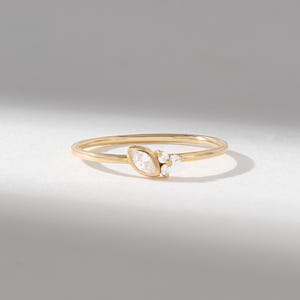 May include: A thin gold ring featuring a marquise-cut clear gemstone set horizontally, accompanied by three smaller round-cut clear gemstones clustered to the side. The band is smooth and polished, showcasing a delicate and refined metal construction.