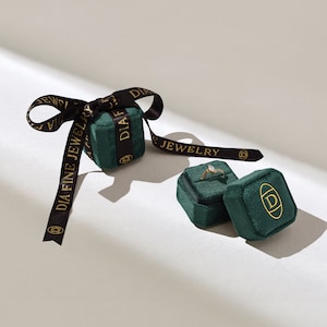 以下が含まれることがあります： エメラルドグリーンのベルベットジュエリーボックスのコレクション。1つは「DIA FINE JEWELRY」と金色のプリントが施された黒いリボンで結ばれています。1つのボックスが開いており、ダイヤモンドの付いたゴールドのリングが表示されています。ボックスには金のロゴがあります。