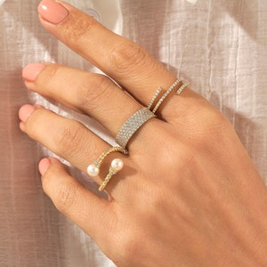 May include: A hand wearing four gold rings. Two of the rings are set with small diamonds, one ring is a twisted band with two pearls, and the other ring is a simple band with a single pearl.