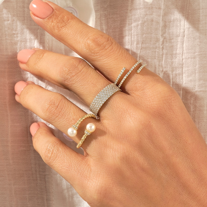 May include: A hand wearing four gold rings. Two of the rings are set with small diamonds, one ring is a twisted band with two pearls, and the other ring is a simple band with a single pearl.